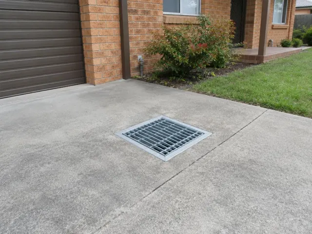 Concrete driveway with a square metal drain grate beside brick house and garage in Bolton Point