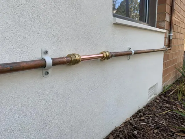 Exterior wall with exposed repaired copper water pipe and wall clips near a window in Woodrising